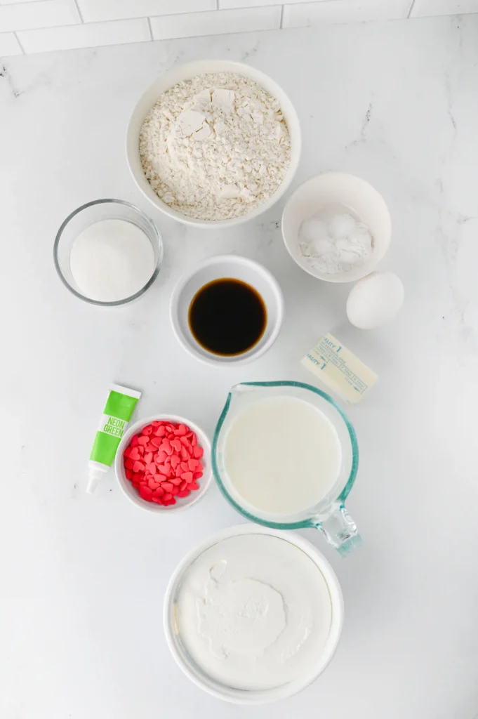 ingredients for the grinch pancakes: flour in a bowl, sugar, vanilla, eggs, milk, pink heart candies, and icing tube on a marble surface.