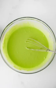 glass bowl filled with green colored pancake mix and a whisk