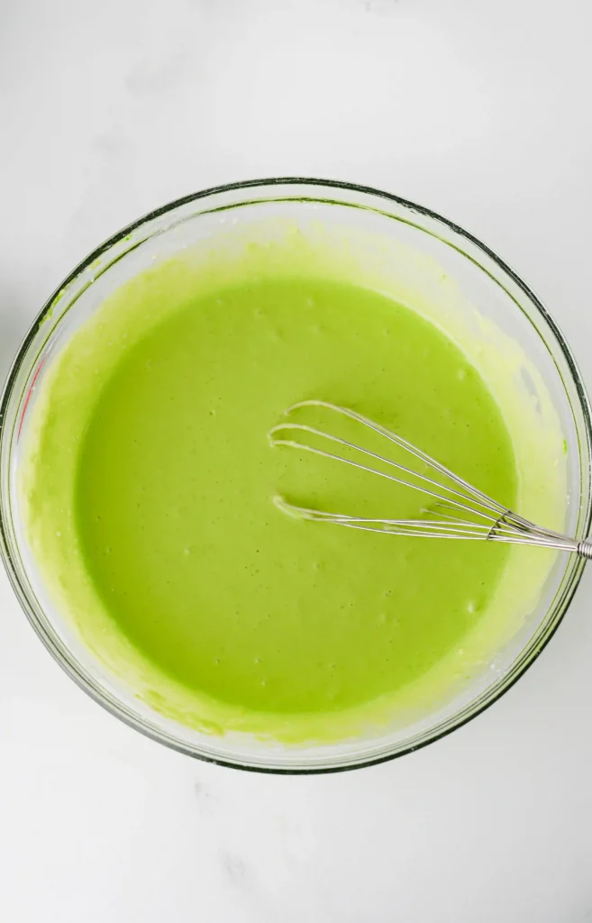 glass bowl filled with green colored pancake mix and a whisk