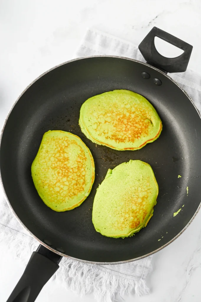 Green pancakes browning in a black nonstick skillet on a light surface, spatula nearby.