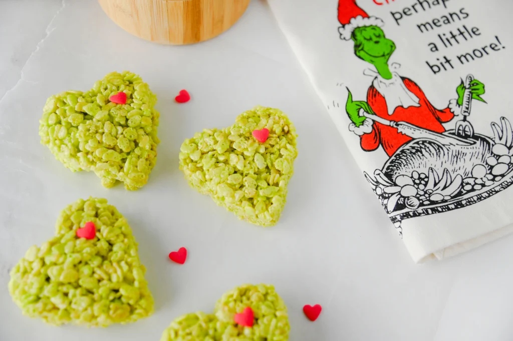 Heart-shaped green rice krispie treats with pink heart sprinkles on a white marble surface with a festive cartoon napkin nearby
