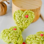 Heart-shaped green rice krispie treats with pink heart sprinkles on a white marble surface with a festive cartoon napkin nearby