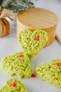 Heart-shaped green rice krispie treats with pink heart sprinkles on a white marble surface with a festive cartoon napkin nearby