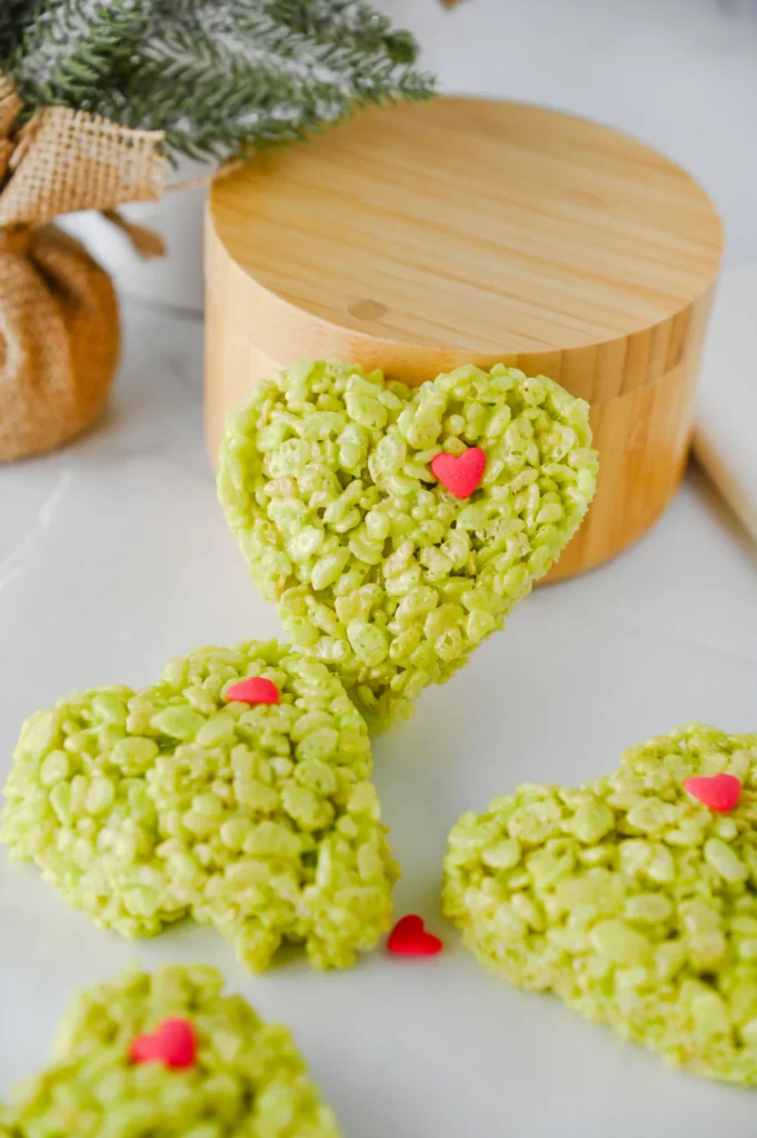 Heart-shaped green rice krispie treats with pink heart sprinkles on a white marble surface with a festive cartoon napkin nearby