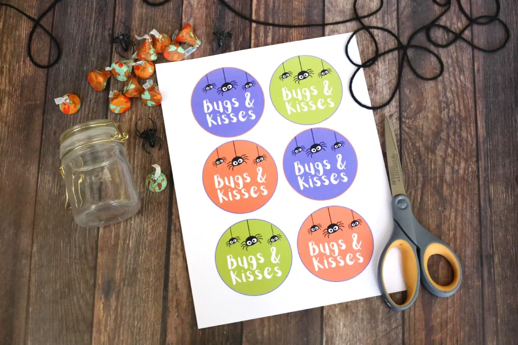supplies for the halloween printable tags: Sheet of round stickers on a white page, each saying'Bugs & Kisses' with cute spider graphics in purple, green, orange, and blue, on a wooden table with scissors nearby.