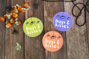 Round colorful tags reading 'Bugs & Kisses' with cartoon spiders, on a rustic wooden surface, next to orange candy wrappers.