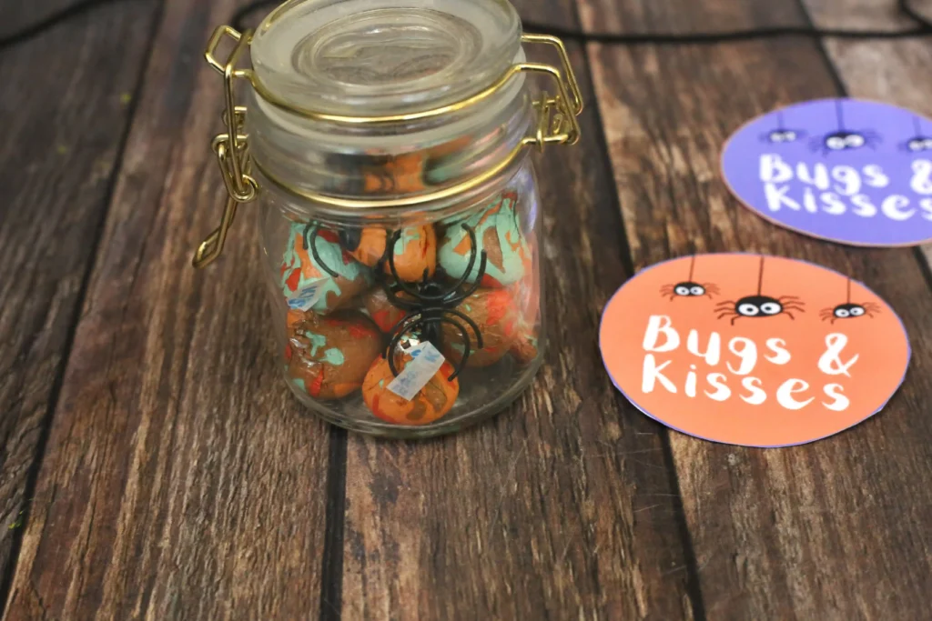 Glass jar filled with orange and turquoise candy and a black spider ornament on a wooden surface, beside colorful'Bugs & Kisses' sticker cards (Halloween candy display).