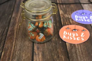 Glass jar filled with orange and turquoise candy and a black spider ornament on a wooden surface, beside colorful 'Bugs & Kisses' sticker cards (Halloween candy display).