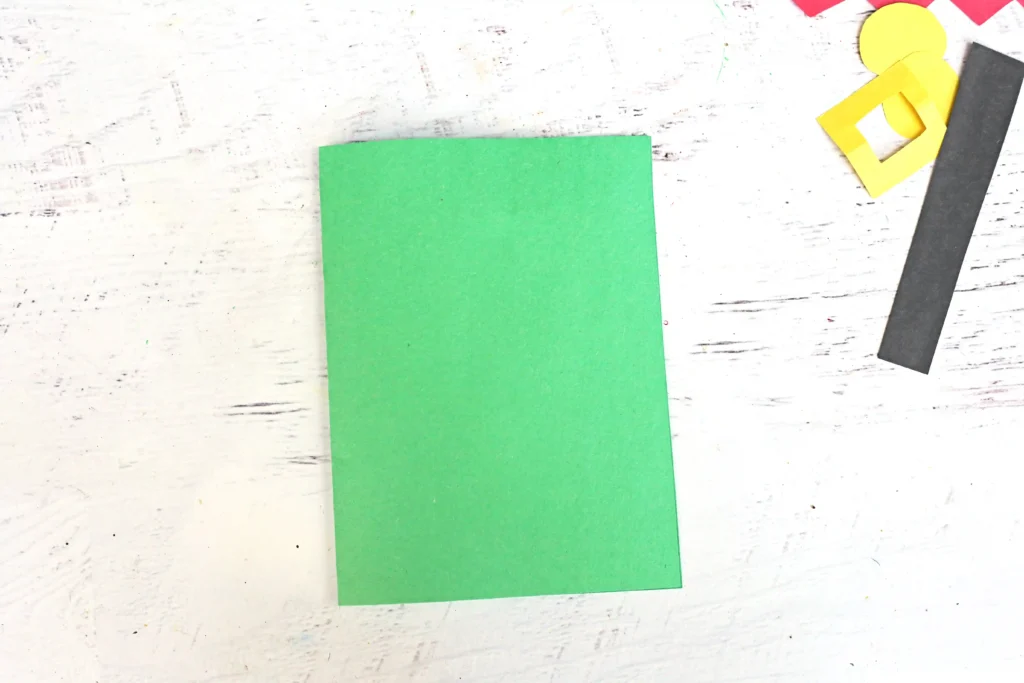 Green square sheet of paper on a light wooden surface with scattered yellow, black, and red craft scraps nearby (arts and crafts supplies).