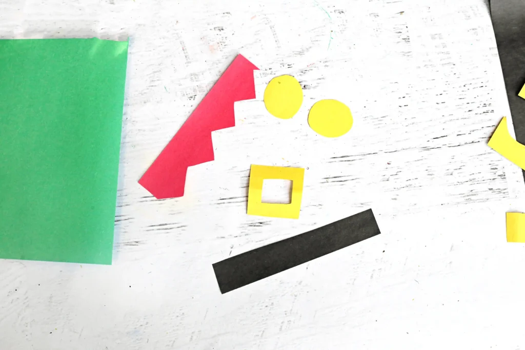 Assorted colorful paper cutouts on a white weathered surface: green rectangle, red zigzag strip, two yellow circles, a yellow square frame, and a black strip.