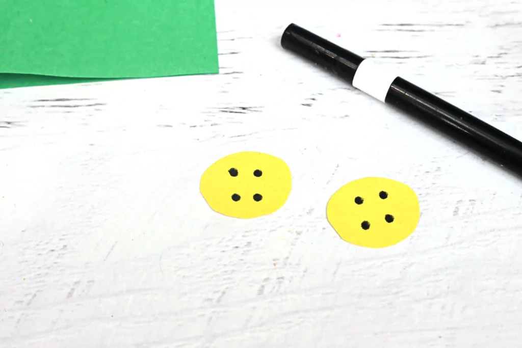 Green folded paper with yellow button-shaped pieces and a black marker on a white wooden surface; crafting scene.