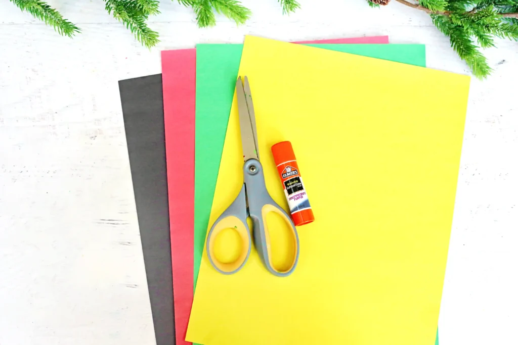 supplies for the elf christmas card: Assorted colored craft paper sheets (black, red, green, yellow) with scissors and glue on a white wooden surface; evergreen branches frame the top.