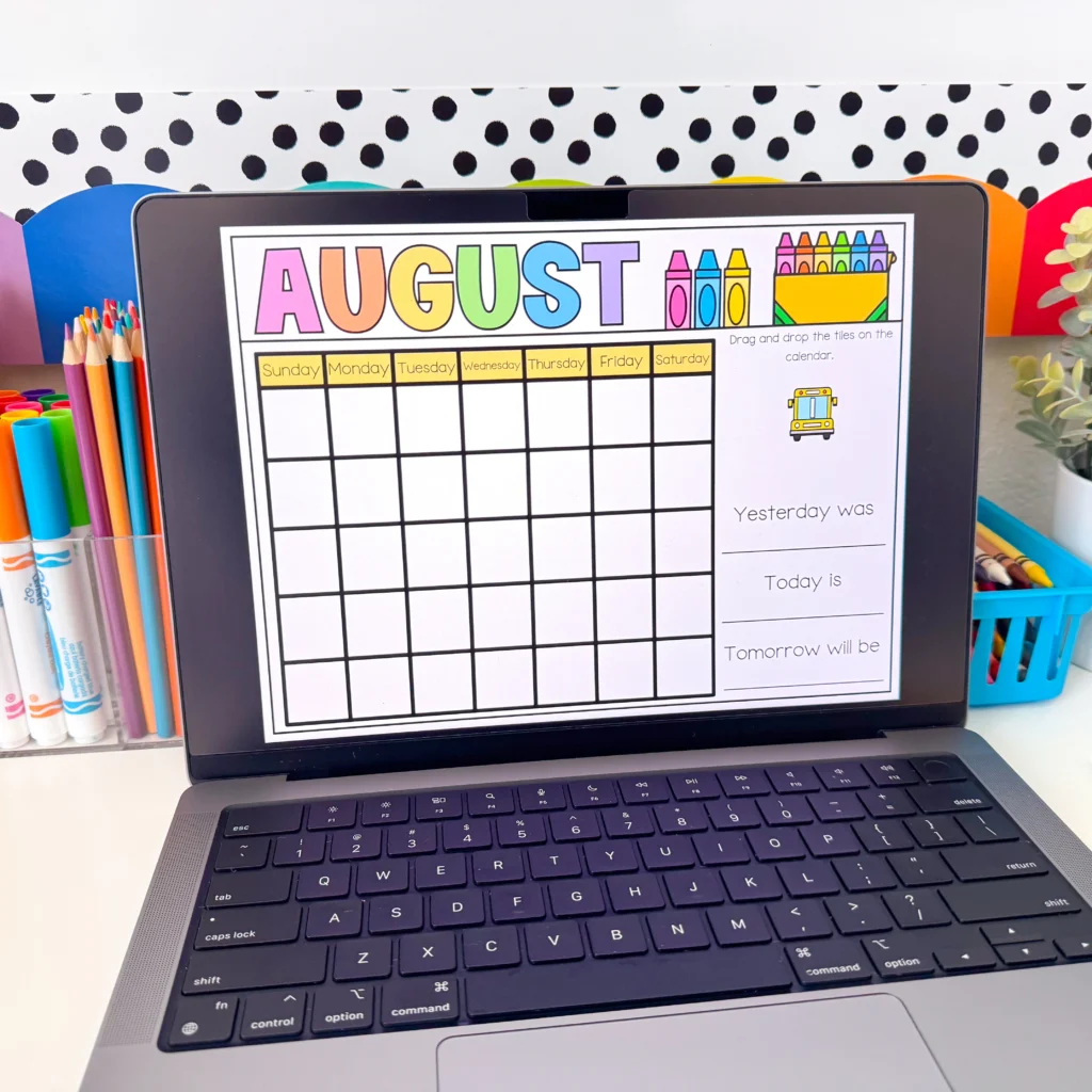 Calendar dashboard on a laptop screen showing the word AUGUST in colorful letters with a row of colored pencils on the left and classroom decor in the background.