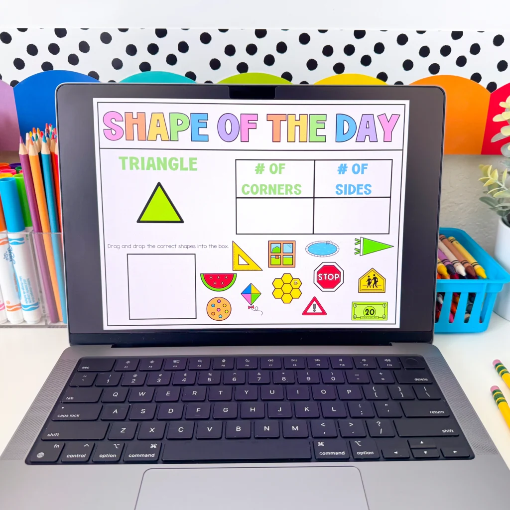 Laptop screen displaying a colorful'Shape of the Day' worksheet with a bright yellow triangle and shape charts beside colored pencils on the desk.