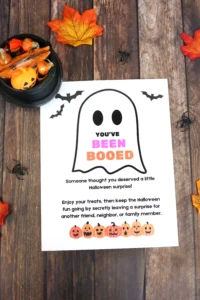Halloween-themed flyer with a ghost outline and the phrase 'YOU'VE BEEN BOOED' in colorful letters, surrounded by pumpkins and bats on a wooden surface.