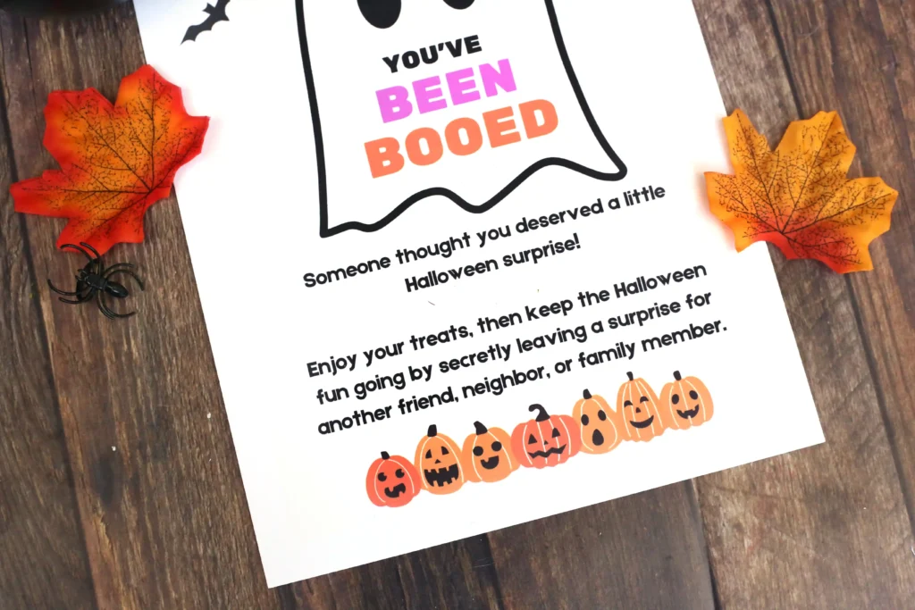Halloween-themed flyer reading "YOU'VE BEEN BOOED" with pumpkin border on a wooden table and autumn leaves nearby.