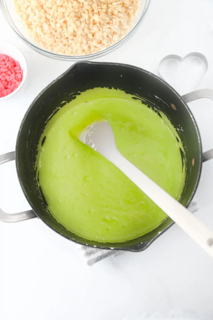 Pot with bright green melted marshmallows and a white spatula, with a bowl of puffed rice and a pink candy heart container in the background.