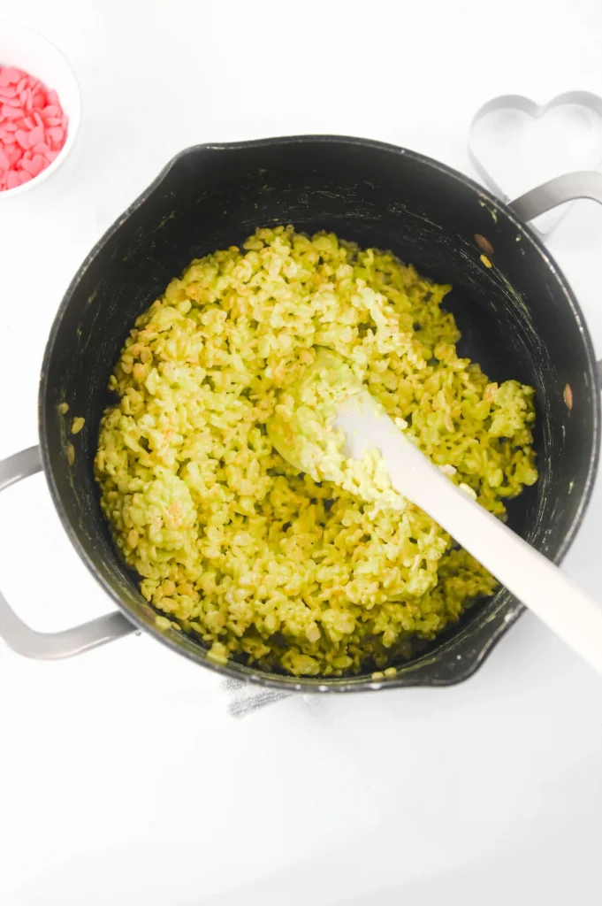 Pot of green rice krispies being stirred with a white spatula, on a white countertop; a bowl of pink heart candies sits nearby.