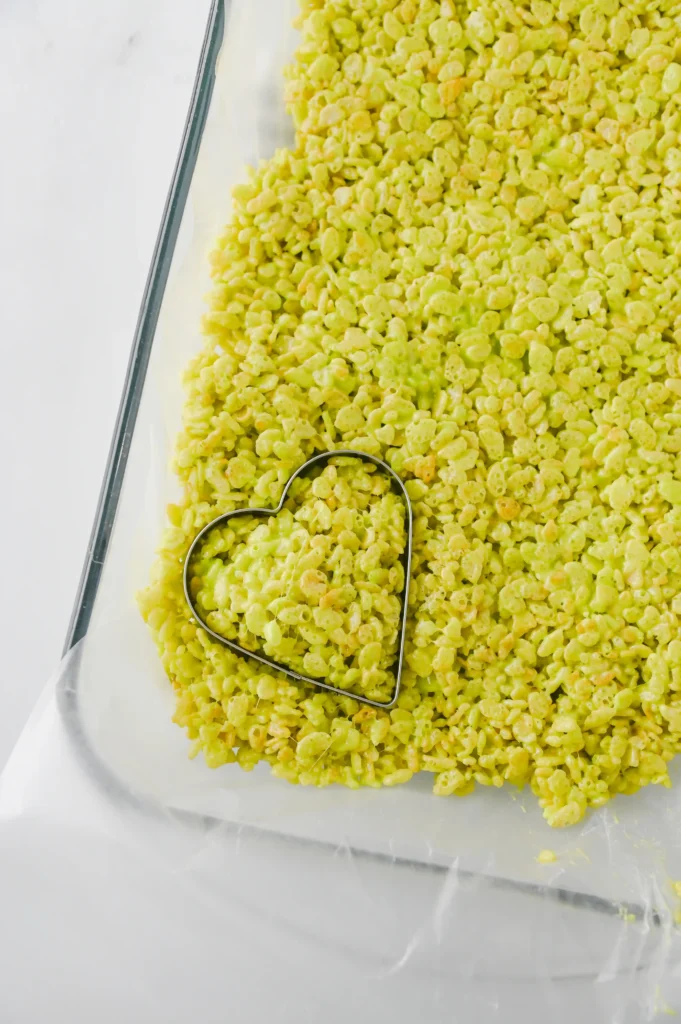 Close-up of a glass dish filled with green puffed cereal, with a heart-shaped metal cookie cutter resting among the pieces.
