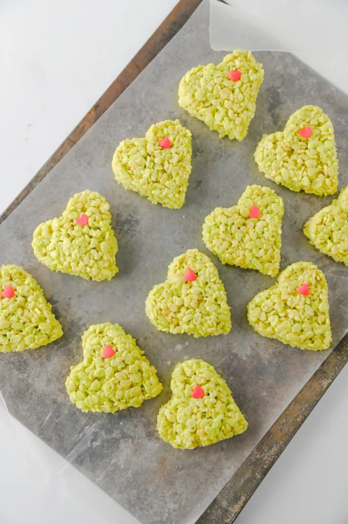 Heart-shaped green rice krispie treats with pink heart sprinkles on a white marble surface with a festive cartoon napkin nearby