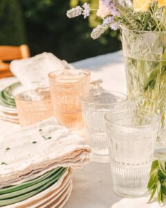 Outdoor table setting with clear ribbed glasses, pastel napkins, and stacked plates next to a vase of pale flowers.