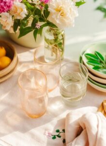 Table setting with a vase of pink and cream flowers, pink-tinted glassware, a clear glass of water, a basket of lemons, and green-rimmed dishes on a pale tablecloth.