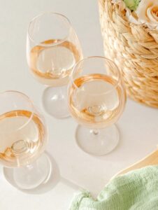Three stemware wine glasses with rosé on a light table, near a woven basket of flowers and a green napkin.