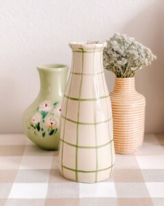 Three ceramic vases sit on a light checkered tablecloth; a tall white vase with a green grid pattern stands at the front, flanked by a green vase with white flowers and an orange-striped vase with dried blooms nearby.